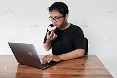 年轻的亚洲人正在吃甜甜圈和喝咖啡，同时使用笔记本电脑工作。印度尼西亚男子身穿黑色衬衫孤立灰色背景。