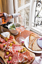 person pouring beverage on clear long-stem wine glass beside white ceramic plate with sliced bread and brown wooden board with bacons