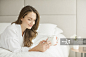Smiling woman in bathrobe drinking coffee and texting on cell phone on bed_创意图片
