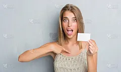 美丽的年轻女子越过肮脏的灰色墙壁拿着空白拜访卡与惊讶的脸指向自己的手指