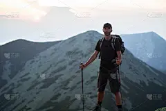 一名白种人徒步旅行者背着背包，拄着拐杖，站在绿石山的山顶上，在夕阳的余晖中，一览群山的全景