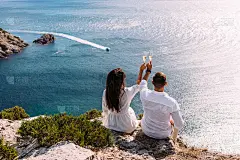 一对相爱的情侣在海边喝着香槟，背对着镜头。这对新婚夫妇在庆祝订婚。一对男女背对着镜头坐在海边喝香槟