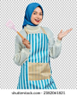 Cheerful beautiful Asian muslim woman wearing apron holding spatula and spreading hands doing housework isolated over purple background. Housekeeping concept 库存照片