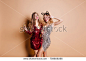 Pale girl in party hair accessory making funny faces for photo. Indoor portrait of gorgeous caucasian woman in red outfit posing with wine goblet, embracing friend.