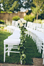 White ceremony chairs along the aisle were adorned with garlands of greenery embellished with soft, ivory roses. #weddingceremony #decor Photography: David & Tammy Molnar. Read More: https://www.insideweddings.com/weddings/an-exquisite-backyard-weddin