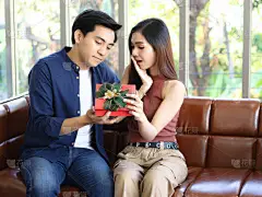 一对年轻的亚洲夫妇坐在客厅的沙发上，彼此愉快地看着对方，而女人则打开一个男人送给她的礼品盒.