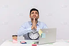 一名不开心的员工坐在办公室工作场所，拿着钟看着恳求用祈祷的手势，乞求