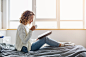 Beautiful young woman sitting on bed in morning, reading book, wearing white knitted sweater, drinking coffee