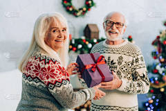 一位快乐的老妇人一边看着相机，一边给快乐的丈夫送圣诞礼物