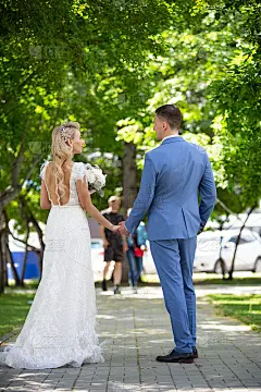 年轻美丽的一对新婚夫妇，新郎和新娘在他们的婚礼当天手牵手走在绿树环绕的公园里。夫妻在成家之前的爱情和