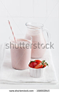 Glass of delicious strawberry smoothie with pink straw served on white linen on top of white table with a pitcher and cup of fresh strawberries.  - stock photo