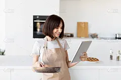 快乐的年轻家庭主妇在厨房做饭，穿着围裙，拿着煎锅，通过电子平板电脑浏览食谱