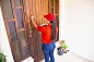 Latin courier knocking on door, holding tablet and carton boxes. brunette long-haired deliverywoman in red uniform standing in front of door and delivering order. delivery service and post concept