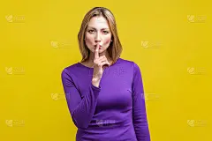 嘘,安静点!肖像严肃的女人在紧紫色连衣裙显示沉默的手势。孤立在黄色背景上