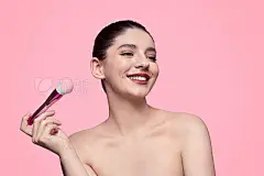 Smiling model with makeup brushCheerful young woman with red lips smiling and looking away while holding soft makeup brush against pink background