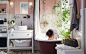A man sits in the bath in a bathroom with pale pink walls, white tiles, white sink unit and plants.