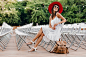 Attractive woman dressed in white dress, red hat, sunglasses sitting in summer open air theater on chair alone, spring street style fashion trend, accessories, backpack, social distancing