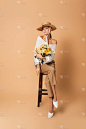 全长的年轻女子头戴草帽，手里拿着一束鲜花，坐在米黄色的木制凳子上  