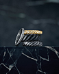 A color photograph shows an 18K white gold wedding band leaning against a stack of three wedding bands in 18K yellow gold and black or grey titanium. Each ring is from the Cable Collection. The rings sit atop a dark, marbled stone surface with a dark bac