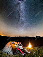 幸福的情侣游客期待着普照满天星斗的天空，坐在靠近发光帐篷和晚上露营篝火晚会。天文摄影