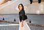 Attractive stylish woman walking in street in fashionable outfit, holding suede handbag, wearing black leather jacket and white lace dress, spring autumn style, turning around in motion