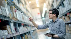 年轻的亚洲工人正在用数码板和笔清点仓库货架上的纸箱里的产品。实物盘点概念