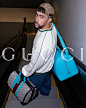 Bad Bunny, a Latino man with a beard and a backwards beige and brown GG monogram cap turns around as he goes down an escalator. He is wearing a white sweatshirt with dark green trim, baggy blue jeans. Over his shoulder is a neon blue duffle. In his hand i