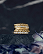 A color photograph shows a stack of five 18K yellow gold wedding bands with or without diamonds from the DY Unity and DY Eden collection. The stack of rings sits atop a dark, marbled stone surface with a dark background and purple flowers in the foregrou