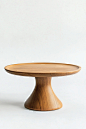 A simple wooden cake stand with a wide base and low profile, designed in the style of William Morris for Urban Outfitters. Product photography on a white background, straight-on view.