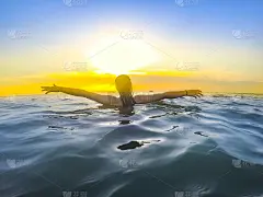 年轻女子举起双手，站在海水中，望着地平线上的夕阳