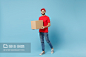 穿着红色制服的送货员隔离在蓝色背景,Delivery man in red uniform isolated on blue background, studio portrait. Male employee in cap t-shirt print working as courier dealer hold empty cardboard box. Service concept. Mock up copy space. - 图虫创意-全球领先正版素材库-Adobe Stock中国独家合作伙伴