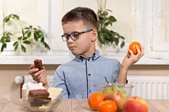巧克力棒或橘子。一个学龄男孩坐在桌旁，不知道该选什么。