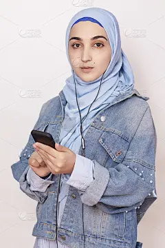 现代穆斯林女孩在智能手机上听音乐、播客和有声读物。快乐的穆斯林妇女戴着头巾听着歌曲
