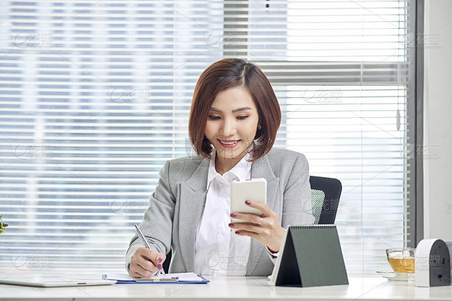 女商人在办公室边看手机边做笔记。女企业家坐在桌子边写笔记边玩手机。素材-花瓣网