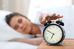 昏昏欲睡的年轻女子伸出手来敲响警钟，愿意关掉它。早醒、睡眠不足的概念