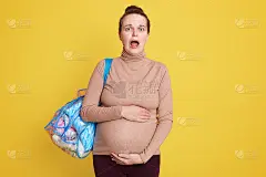 震惊的准妈妈在黄色背景下摆出孤立的姿势，大张着嘴，非常害怕去产科之家，双手一直放在肚子上。