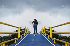 一个年轻的女人跑上一座小山，在一座运河上的人行天桥上，一条蓝色的柏油路，一条黄色的护栏。