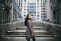 一个商业主题的女人的肖像。年轻的白人黑发女孩穿着长夹克，外套和黑色皮包站在商业中心的背景，办公大楼的