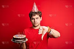 年轻的卷发金发男子戴着生日帽，拿着蛋糕盖在红色背景上，愤怒的脸，表示不喜欢，拇指向下，拒绝的概念