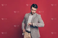 一个守时的，刚强的，严厉的，惊人的金融家看着他手臂上的手表，紧张他的伴侣迟到，孤立在红色背景，检查时