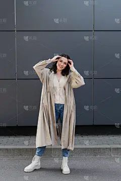 穿着长雨衣和白靴的微笑女人在灰墙附近理发