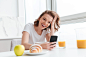 Portrait of laughing woman in white tshirt checking news on mobile phone while sitting at the kitchen table