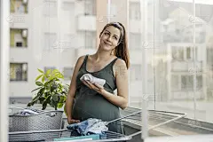 母性本能，家务和孕妇。一位兴奋的准妈妈在家里的阳台上为新生儿准备衣服并摆姿势拍照。亲情母爱母爱，温情