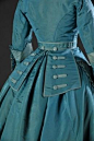 Back view, Dress in turquoise and light blue silk taffeta, 1865-69. This dress in turquoise and pale blue taffeta silk has a train and a separate waistband with panels at the back, with large decorative buttons. The daytime corsage has a V-shaped neckline