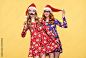 Christmas New Year. Young Woman in Santa Claus hat Having Fun Happy with Holiday Props. Fashion. Pretty Playful Sisters Best Friends.Twins in Stylish fashion Red XmasDress on Yellow.Christmas Colorful