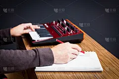 在播放音乐键盘合成器的同时，靠近正在白纸上写着热门歌曲的男性作曲家的手。写歌的概念