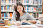 亚洲青年学生穿着休闲服在大学或学院的图书馆里阅读和做作业，书架背景上方的木桌上放着各种书籍和文具，回