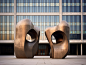 Henry Moore's sculpture features abstract shapes made of bronze material standing in front of the building. It has a symmetrical composition with large glass windows on both sides. The background features gray concrete walls with strong light reflection e