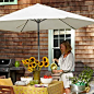 woman standing on a bright patio behind a shingled house pouring from a yellow pitcher over a decorated and set table covered in the shade of a large white patio umbrella, with a grill in the background