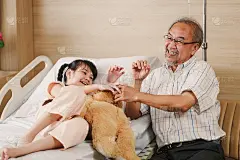 可爱的亚洲小女孩坐在医院的床上和她的爷爷一起读书。生病的女孩抱着泰迪熊和她的爷爷度过了快乐的时光。儿